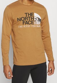 Langärmliges, braunes Baumwoll-T-Shirt mit einem grafischen Print, der einen Bergdesign und schwarz-weißen Text auf der Vorderseite zeigt.