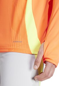 Oranje sporttop met gaasoppervlakken in helder geel en een gestructureerde afwerking. Het label "AEROREADY" is zichtbaar nabij de naad.