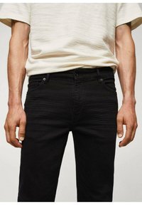 Jean en denim noir avec une coupe ajustée, présentant des poches avant, un bouton à l'avant et une texture lisse. Porté avec un haut à manches courtes de couleur claire.