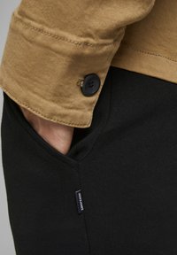 Camicia in corduroy marrone con un bottone nero e felpa nera con tasca laterale, dotata di un piccolo etichetta del marchio.