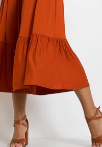 Rdzaworuda spódnica z rozkloszowanym dołem, wykonana z miękkiego materiału, w połączeniu z brązowymi sandałami na wysokim obcasie z paskami.