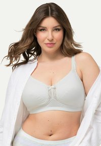 Soutien-gorge blanc avec des détails en dentelle au centre, bretelles ajustables et tissu doux. Le modèle porte une chemise blanche par-dessus. Texture lisse.