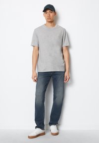Tricou din bumbac gri, cu mâneci scurte, asociat cu blugi denim de culoare albastru închis. Modelul poartă o șapcă bleumarin și adidași albi cu talpă din cauciuc.