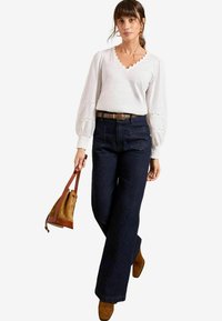 Top bianco a maniche lunghe con scollatura a scallop e dettagli ad occhio, abbinato a jeans a gamba larga blu scuro a vita alta e cintura marrone.
