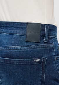 Los pantalones vaqueros de mezclilla azul oscuro presentan una superficie texturizada, un parche de cuero con la marca en la cinturilla y un pequeño logotipo bordado encima del bolsillo trasero.