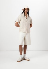 Camisa de manga corta beige claro con dos bolsillos en el pecho, combinada con pantalones cortos beige claros hasta las rodillas. Zapatillas blancas con detalles negros.