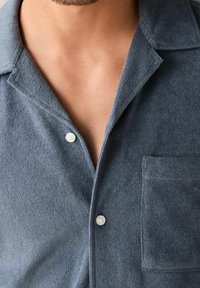 Sinapintainen sininen paita, jossa kaulus, yksittäinen rintatasku ja valkoiset napit. Kangas näyttää pehmeältä ja hieman pörröiseltä.