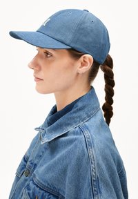 Blauer Denim-Baseball-Cap mit gebogenem Schirm und gesticktem Logo. Kombiniert mit einer Jeansjacke und einem Hemd mit Kragen darunter.
