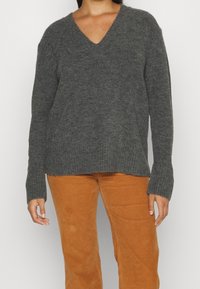 Šedý svetr s V výstřihem vyrobený z měkké pleteniny, s ribbovanými manžetami a lemem. Model jej nosí s béžovými kordovými kalhotami.