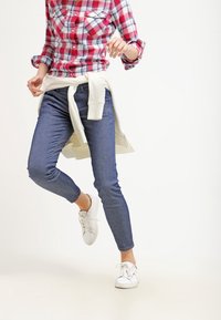 Rutig flanellskjorta i röd och vit, bärs över ljusblå jeans. Vita sneakers. Lätt tröja knuten runt midjan.