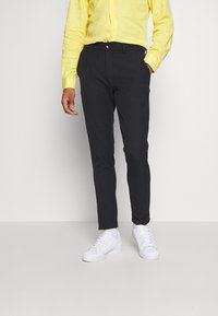 Donkergrijze slim-fit broek met een subtiel patroon, gecombineerd met een lichtgele overhemd met knopen en witte sneakers.