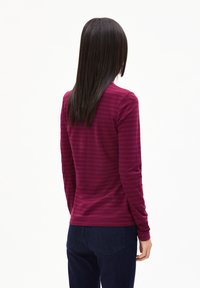 Person mit langen dunklen Haaren, der ein burgunderfarbenes gestreiftes Langarmshirt und dunkelblaue Jeans trägt, von hinten vor einem weißen Hintergrund gezeigt.