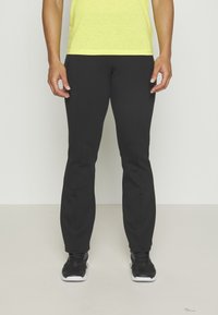 Schwarze Sporthose im Slim-Fit-Schnitt, mit einer glatten Textur und dezenten Nähten. Kombiniert mit schwarzen Laufschuhen und einem leuchtend gelben Shirt.