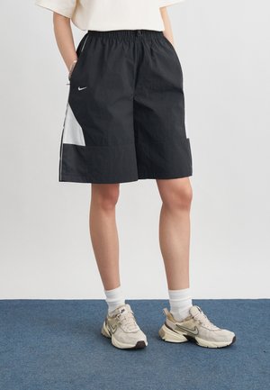 Personne portant un short Nike noir et blanc, des chaussettes blanches montantes et des baskets Nike beige, debout sur un tapis bleu devant un fond blanc.