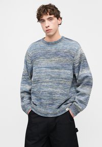 Meerjarige gebreide trui met tinten blauw en groen, voorzien van een klassieke ronde halslijn en geribbelde boorden. De textuur van de stof zorgt voor diepte.