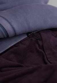 Un hanorac violet și pantaloni din catifea închis violet, cu un model texturat și cordon de strângere, așezate împreună pe o suprafață plană.