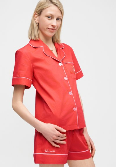 Ξανθιά γυναίκα που φοράει κόκκινο πουκάμισο με κοντά μανίκια και κουμπιά και ταιριαστό σορτς με λευκή περιγραφή, στέκεται μπροστά σε ένα απλό φωτεινό φόντο.