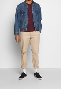 Blaue Jeansjacke mit zwei Brusttaschen über einem bordeauxfarbenen T-Shirt, kombiniert mit khakifarbenen, hochgekrempelten Hosen und schwarzen Sneakers mit weißen Sohlen.