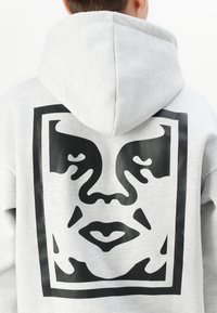 Sweatshirt cinza com uma grande gráfica preta de um rosto emoldurado em um quadrado nas costas. Possui capuz ajustável com cordão e textura de tecido suave.