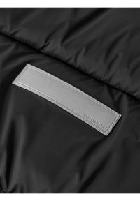 Patch rectangulaire réfléchissant cousu sur une surface en tissu noir matelassé, avec le texte "name it" embossé dans le coin du patch.