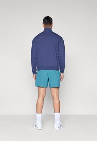 Μπλε ναυτικό φούτερ με ψηλό γιακά, σε συνδυασμό με σορτς σε τιρκουάζ απόχρωση. Το μοντέλο φοράει λευκές κάλτσες και αθλητικά παπούτσια. Απλός και λειτουργικός σχεδιασμός.