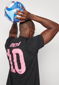 Camiseta negra con el texto "mesl" en rosa y el número "10" impreso en la espalda. Una persona sostiene un balón de fútbol multicolor sobre su cabeza.