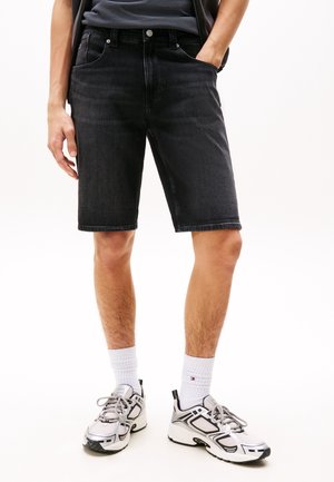 Person iført sorte denimshorts, hvide crew-sokker og hvide og sorte løbesko, der står med den ene hånd i lommen.