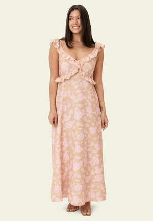 Vrouw draagt een beige mouwloze maxijurk met roze bloemenpatroon en gerimpelde halslijn, staat blootsvoets op een lichte achtergrond.
