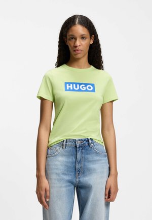 Frau mit langen lockigen Haaren, die ein hellgrünes T-Shirt mit blauem "HUGO"-Logo und hellblaue hoch taillierte Jeans trägt, steht vor einem einfarbigen Hintergrund.