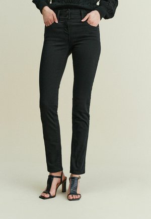 Jeans negros de cintura alta con ajuste slim, que cuentan con cierre de dos botones y bolsillos frontales, combinados con sandalias negras de tacón alto y tiras.