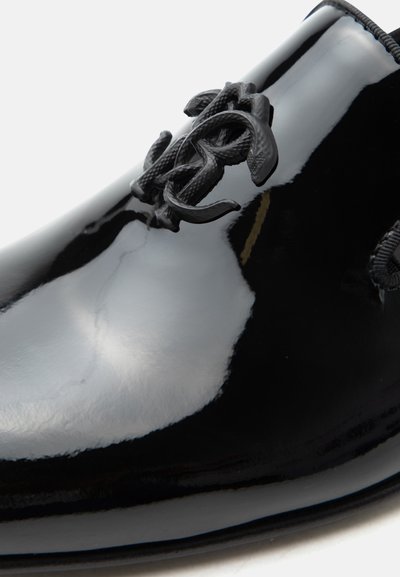 Gros plan sur une chaussure habillée noire brillante montrant des lacets noirs détaillés et des boucles de lacets texturées et décoratives sur la surface supérieure.