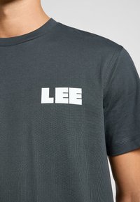 Mörkgrå bomullströja med korta ärmar, med en vit "LEE"-logotyp på bröstet. Ribbad rund halsringning, slät yta och lätt stretch.