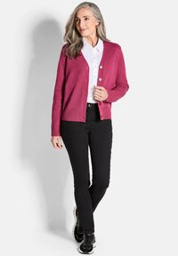 Fuchsia Cardigan mit Knopfverschluss, geripptem Saum, getragen über einem weißen Hemd, kombiniert mit schwarzen Jeans und schwarzen Sneakers. Glatte Textur.