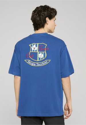 Mand iført blå t-shirt med farverigt broderet "Tennis Club Sergio Tacchini" logo med en stjerne og en tennisbold på ryggen.