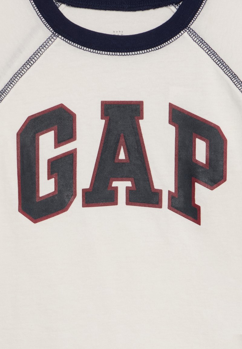 Prenda de tela blanca con un cuello redondo acanalado en color marino. Presenta la palabra "GAP" en grandes letras mayúsculas de color gris oscuro contorneadas en rojo.
