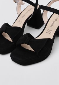 Sandalias de tacón alto de ante negro con tacón de bloque, punta abierta y detalle de lazo. Cuentan con una delgada correa en el tobillo y un forro interior suave.