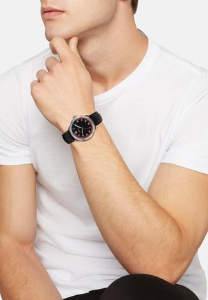 Montre noire avec un boîtier rond, des accents argentés et de grands marquages rouges sur un bracelet noir. Portée sur un poignet clair.