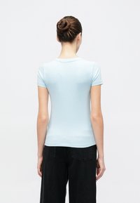 Svetlo modra prilegajoča majica s kratkimi rokavi in okroglim izrezom, pogled od zadaj. Tkanina je videti gladka; v kombinaciji s črnimi širokimi hlačami.