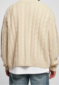 Jersey de punto trenzado en crema, con un dobladillo y puños de canalé, mangas largas y un corte holgado, que presenta patrones de textura intrincados en toda su superficie.