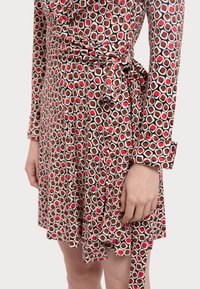 Robe portefeuille à motifs marron et beige avec des accents rouges, manches longues et détail de taille nouée. Tissu lisse à la texture soyeuse.