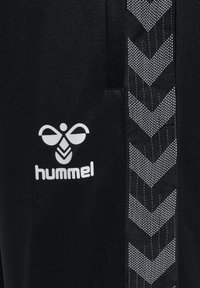 Svarta Hummel träningsbyxor med en vit logotyp, med en strukturerad sidoremsa i fiskbensmönster i grått. Slätt tyg utan fickor.
