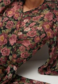 Blomsterbluse med et sort gennemsigtigt stof og pink og creme rosemønstre, fremhævet af en knyttet detalje i taljen.