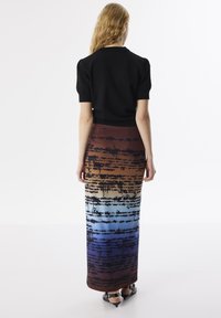 Schwarzes kurzärmliges Oberteil kombiniert mit einem langen, farbverlaufenden Rock in Braun, Orange, Blau und mit Textmustern, dazu glänzende schwarze High Heels.