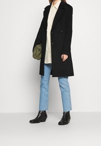 Černý vlněný peacoat s dvojitými knoflíky, přes krémovou košili, doplněný světle modrými rovnými džínami a černými kotníkovými botami na klínu.