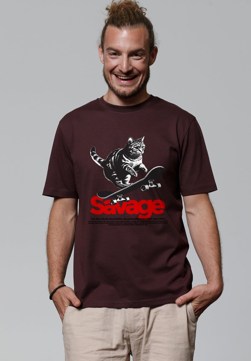 Homme souriant avec les cheveux attachés en chignon, portant une chemise bourgogne ornée d'un chat sur un skateboard et du mot « Savage » en grandes lettres rouges.