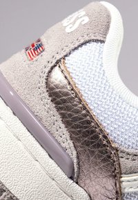 Sneaker con materiale in suede grigio e rete bianca, dettagli metallici, suola testurizzata e una piccola bandiera.