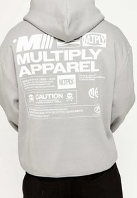 Grå hoodie med vit grafisk text på baksidan, inklusive varumärkesnamn och varningssymboler. Materialet ser mjukt ut; designen är avslappnad och oversize.