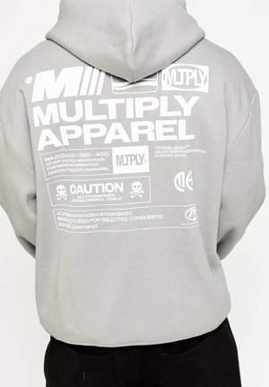 Grijze hoodie met witte grafische tekst op de achterkant, inclusief merknamen en waarschuwingssymbolen. Het materiaal lijkt zacht; het ontwerp is casual en oversized.