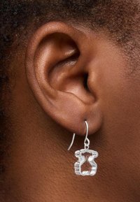 Pendientes en forma de oso plateados con un diseño hueco colgando de un gancho simple, exhibidos contra piel oscura y cabello texturizado cerca de la oreja.