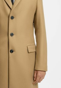 Beige uldfrakke med en glat overflade, tre sorte knapper og en sidelomme. Skreddersyet design med klassisk revers og lange ærmer.